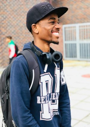 Smiling young man wearing cap and headphones, standing with backpack outdoors.