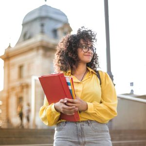 Smiling female student with curly hair and glasses holding books on campus steps.